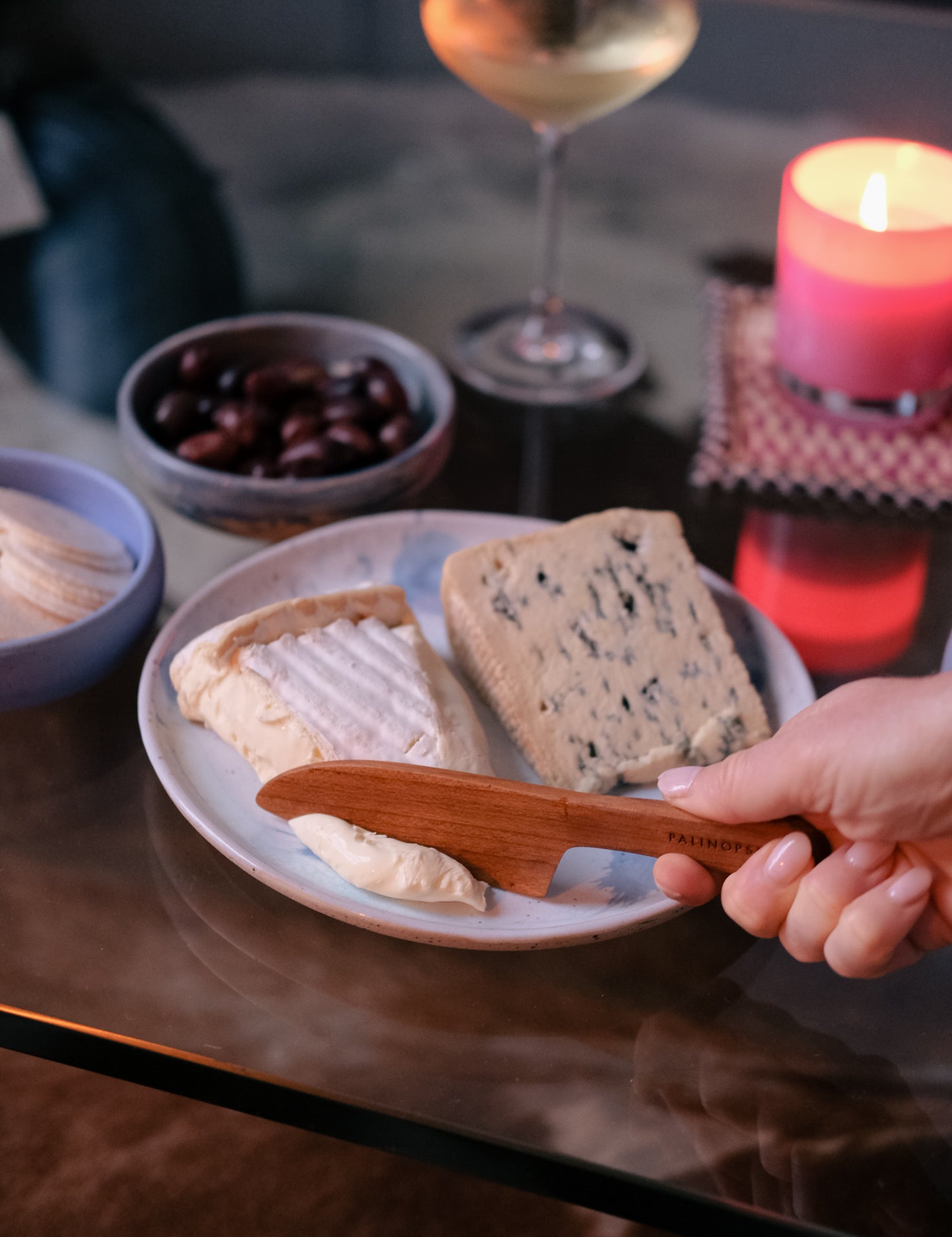 Person cutting soft cheese with a wooden cheese knife, surrounded by a glass of white wine, handmade ceramic bowl of olives, and lit candle on a table.