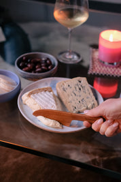 Person cutting soft cheese with a wooden cheese knife, surrounded by a glass of white wine, handmade ceramic bowl of olives, and lit candle on a table.