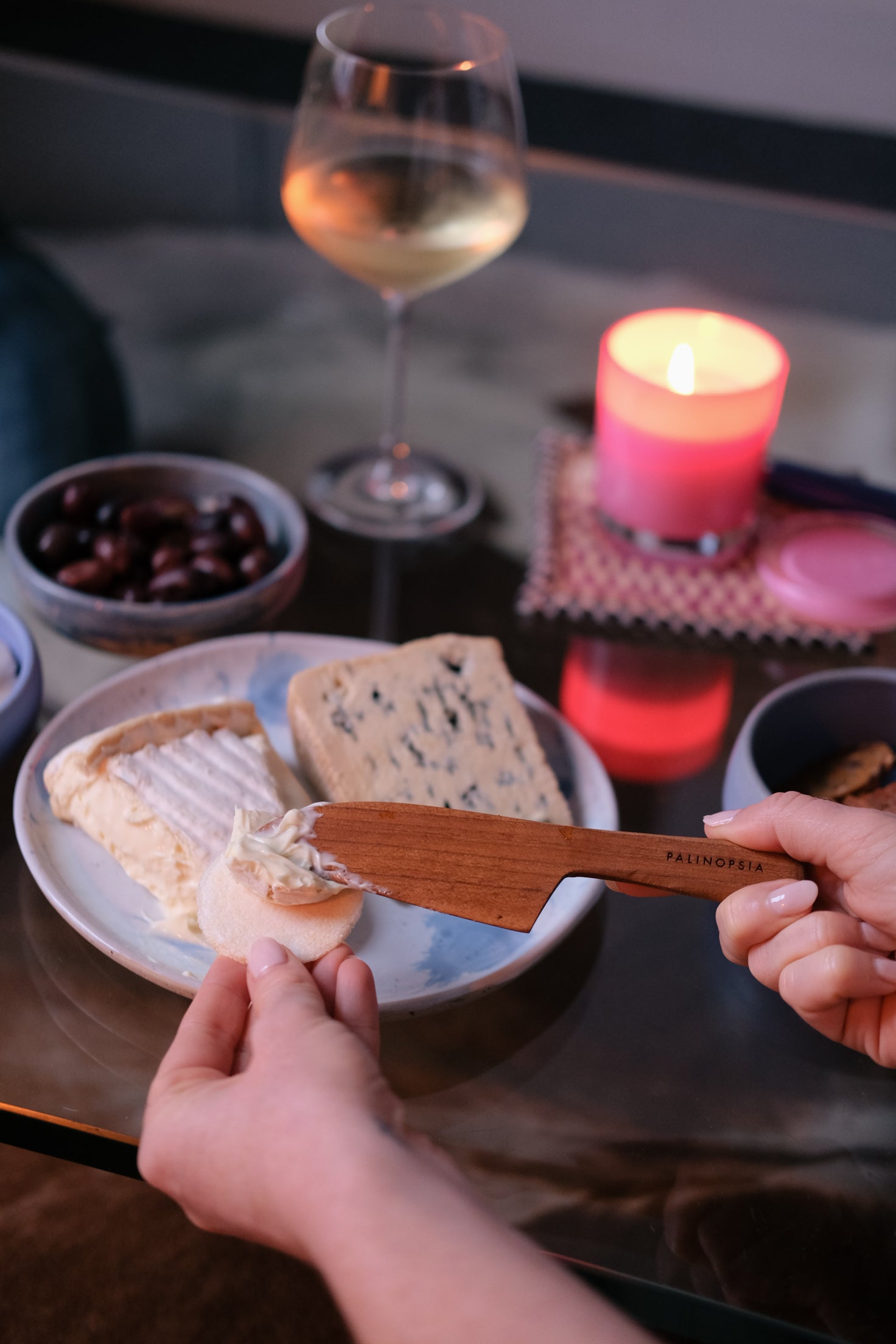 Person spreading soft cheese with a teak wooden cheese knife, surrounded by a glass of white wine, handmade ceramic bowl of olives, and lit candle on a table.