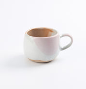 Handmade stoneware ceramic mug with a pinkish hue and chocolate glaze drips on a white background