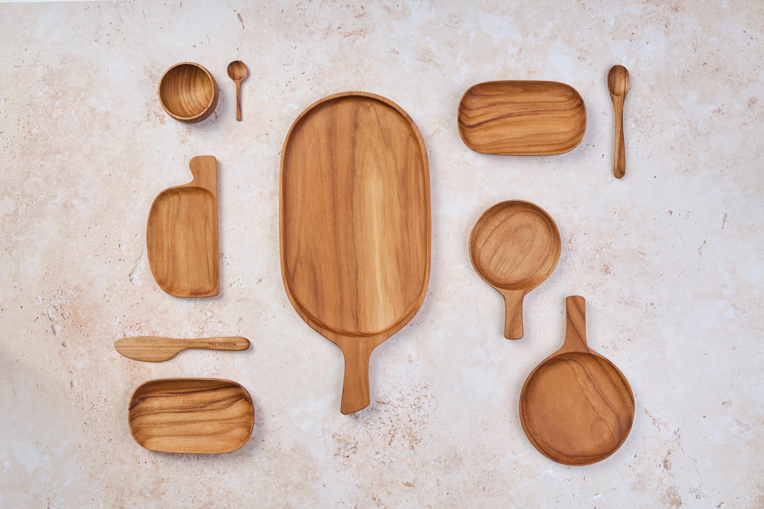 A mix of Palinopsia wooden teak serving trays, small bowls, knives, and cheese boards on a light stone background.