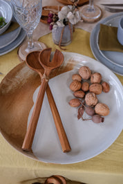 A close-up of a rustic large ceramic serving platter in brown and light blue on a yellow tablecloth, featuring two large wooden salad servers and a cluster of walnuts.