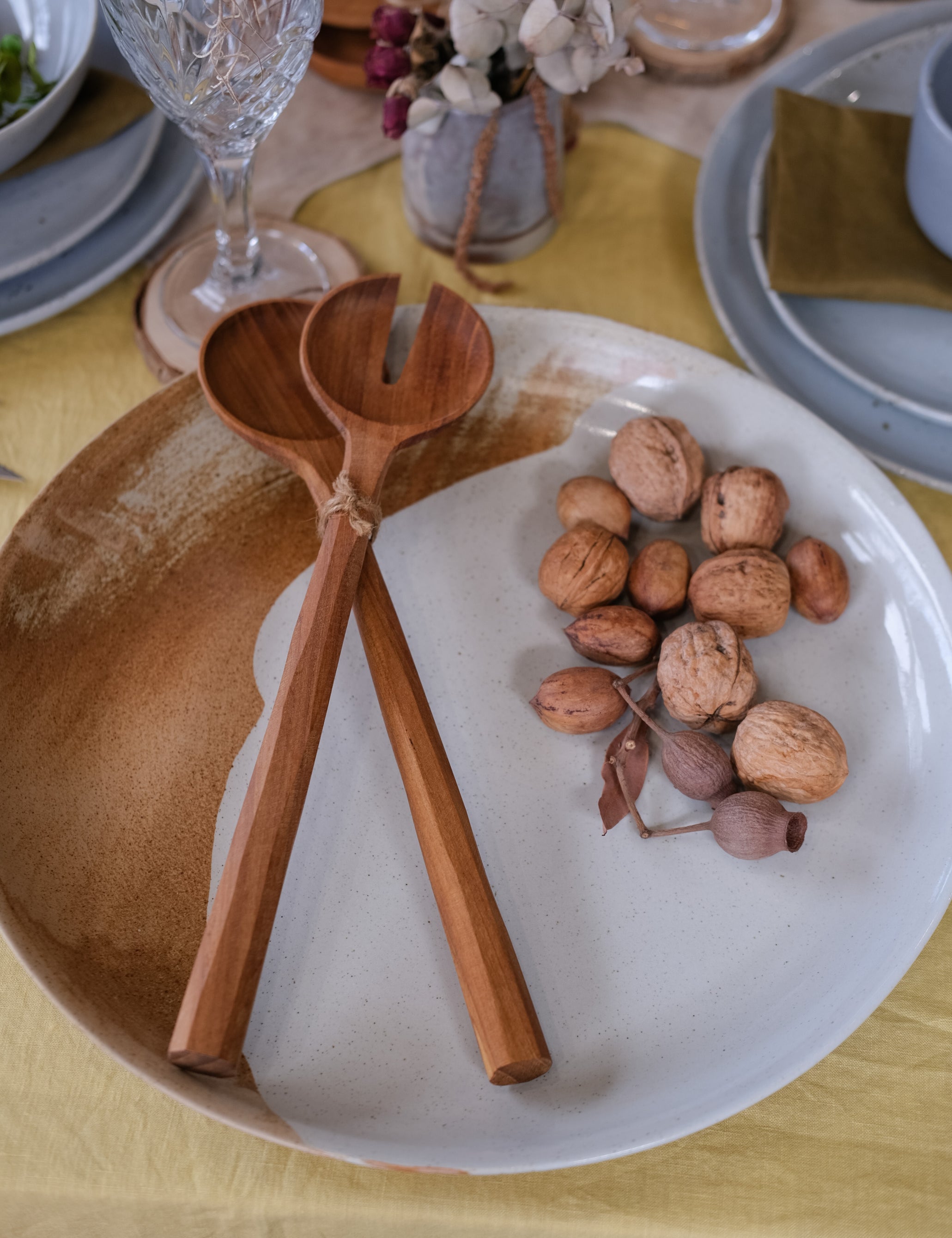 A close-up of a rustic large ceramic serving platter in brown and light blue on a yellow tablecloth, featuring two large wooden salad servers and a cluster of walnuts.