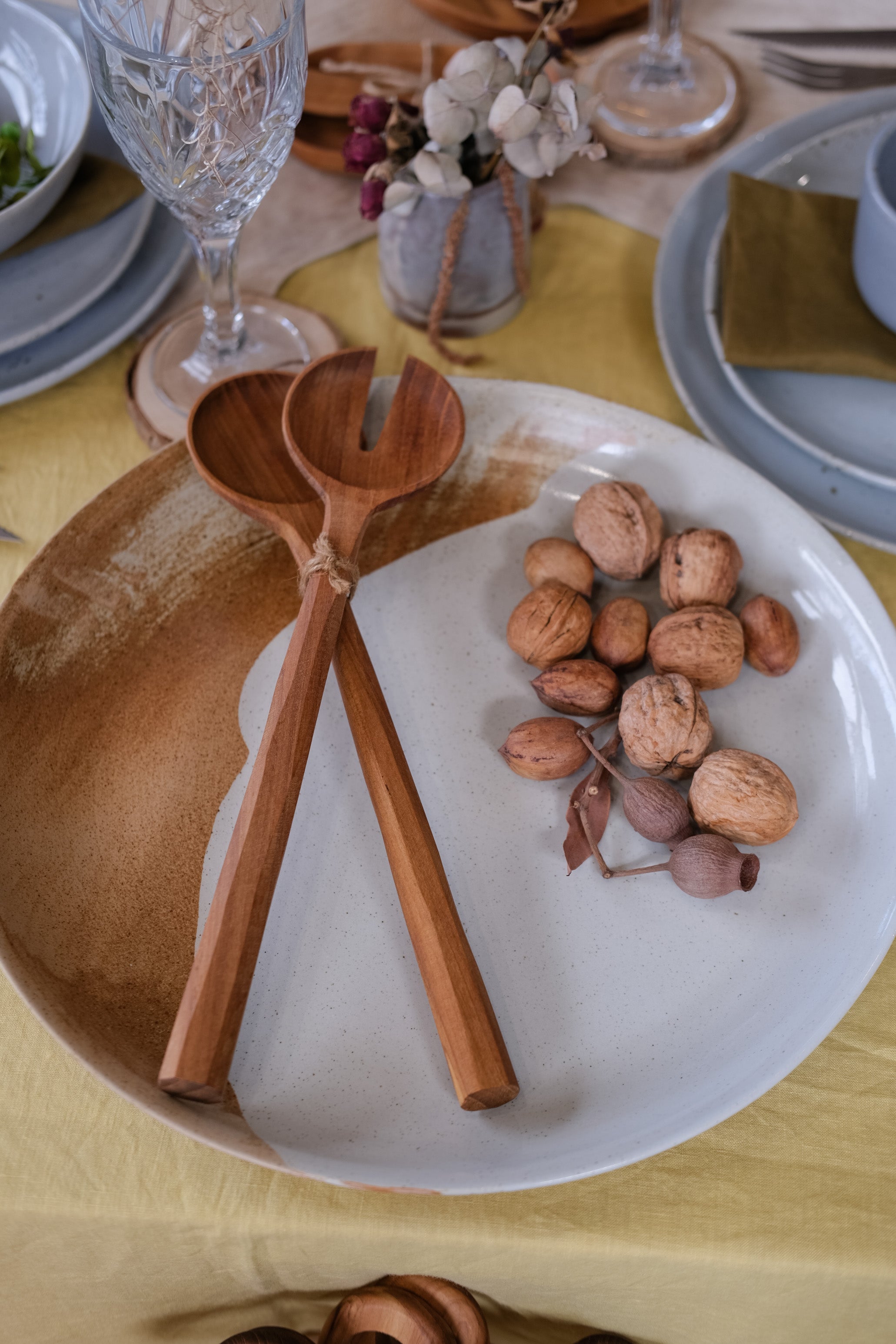 A close-up of a rustic large ceramic serving platter in brown and light blue on a yellow tablecloth, featuring two large wooden salad servers and a cluster of walnuts.