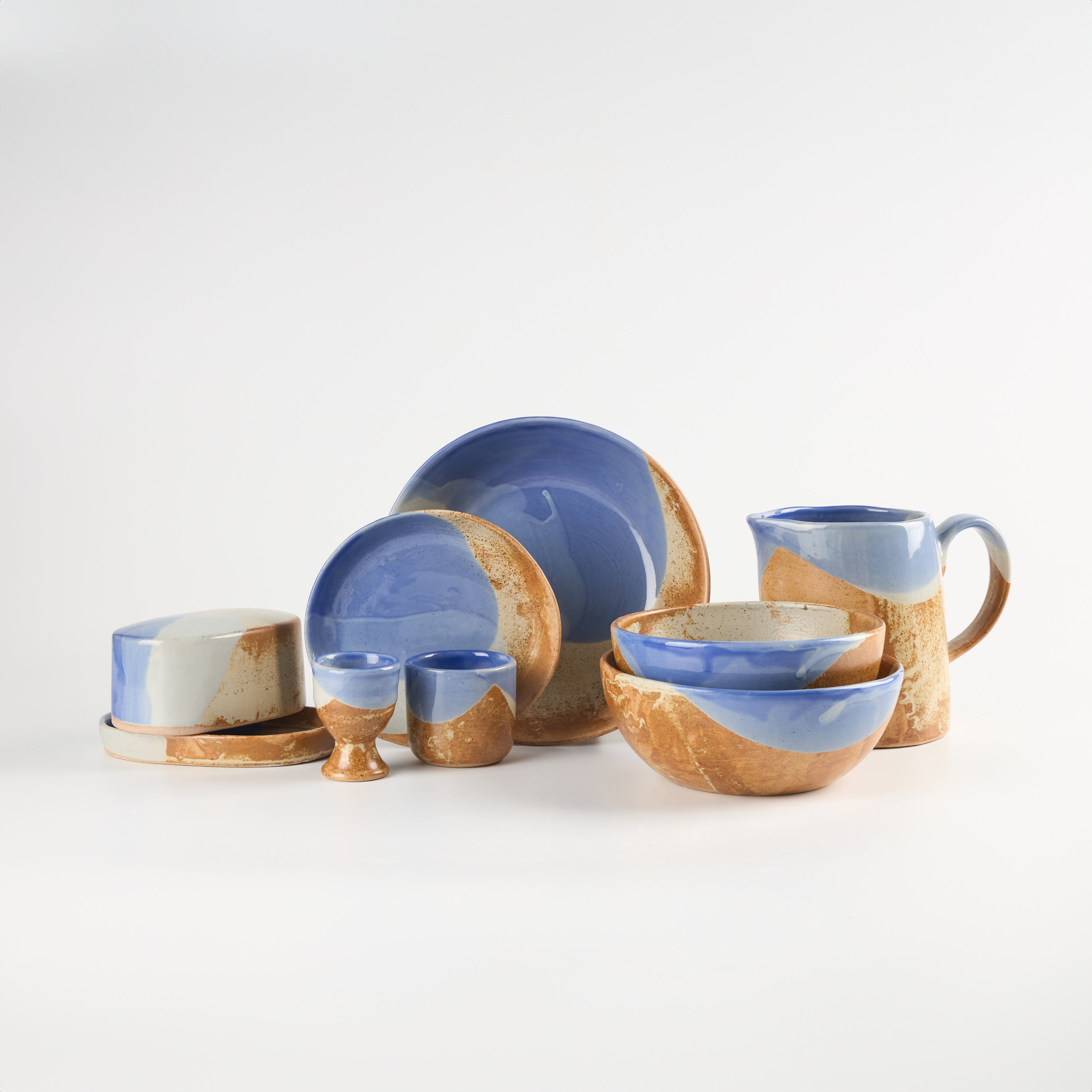 A ceramic tableware set with a glossy cobalt blue glazed finish and a rustic brown contrasting colour, textured tan-brown unglazed edge, displayed on a clean white studio background. Pictured set includes pasta bowls, ramen bowls, egg cups, butter dish, espresso cups and mugs and a large jug. 
