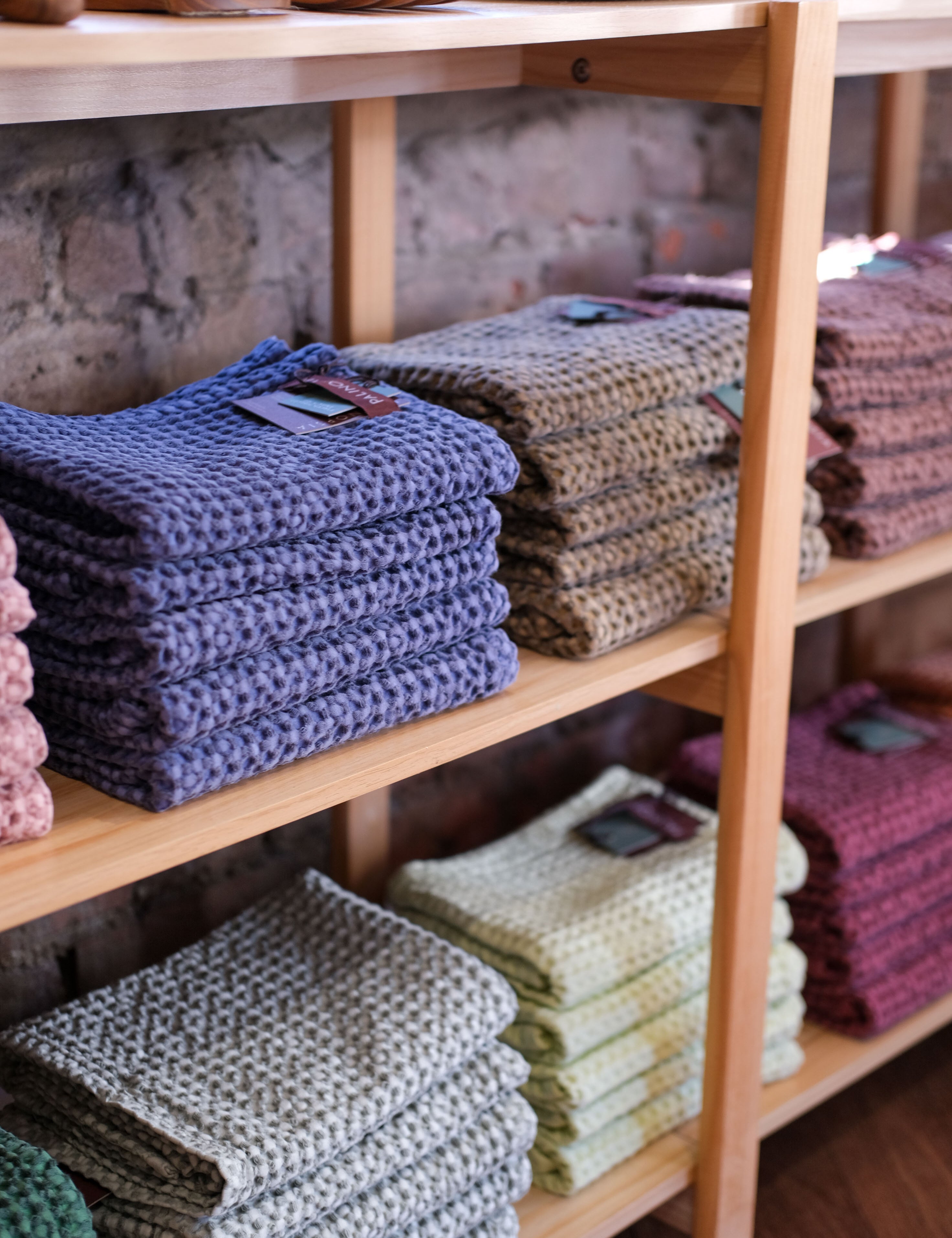 A shelf in Palinopsia’s ceramic store with colourful waffle tea towels folded in stacks, red, light grey, green, denim blue, and pink. On top of the shelf are wooden homewares items. 