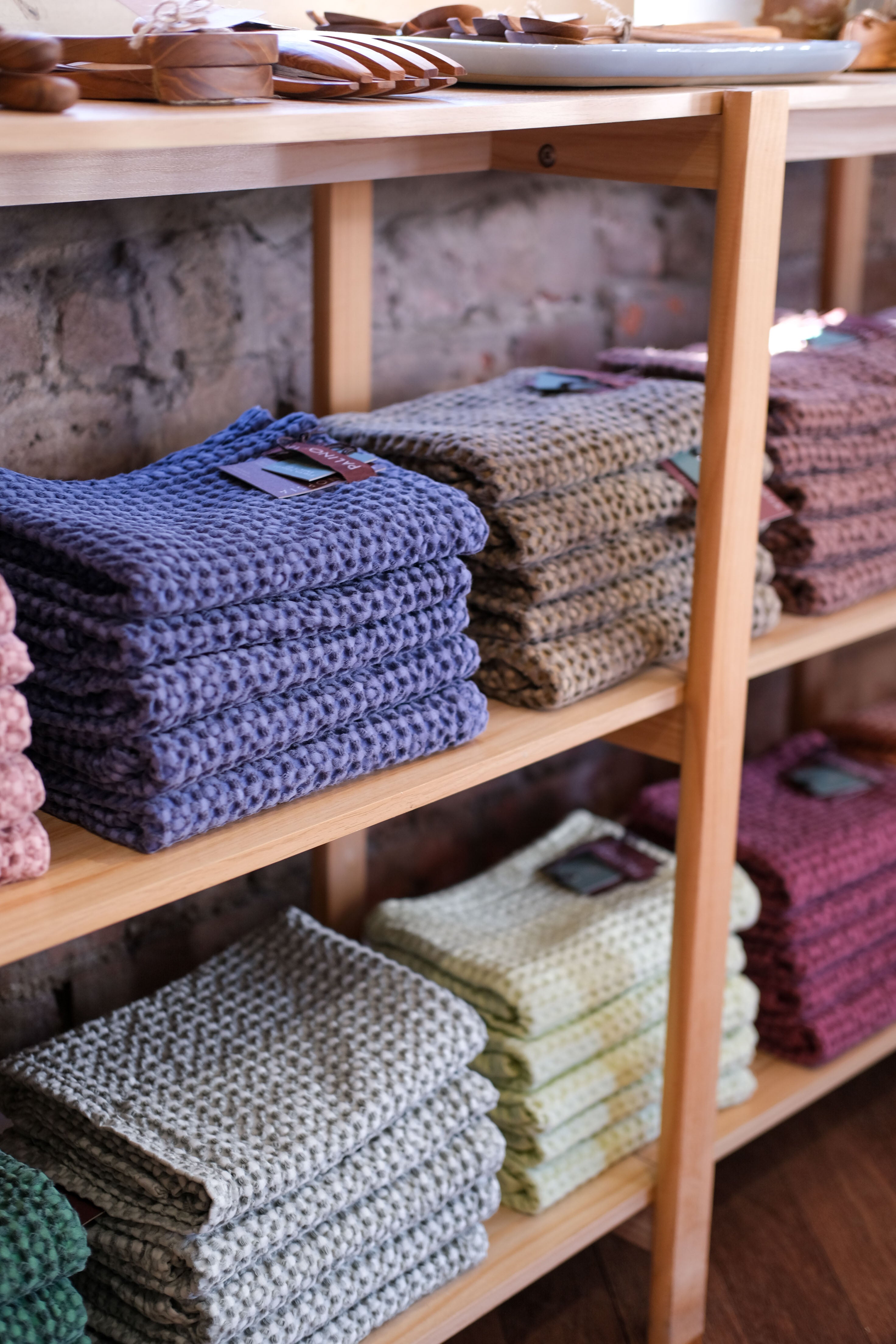 A shelf in Palinopsia’s ceramic store with colourful waffle tea towels folded in stacks, red, light grey, green, denim blue, and pink. On top of the shelf are wooden homewares items. 