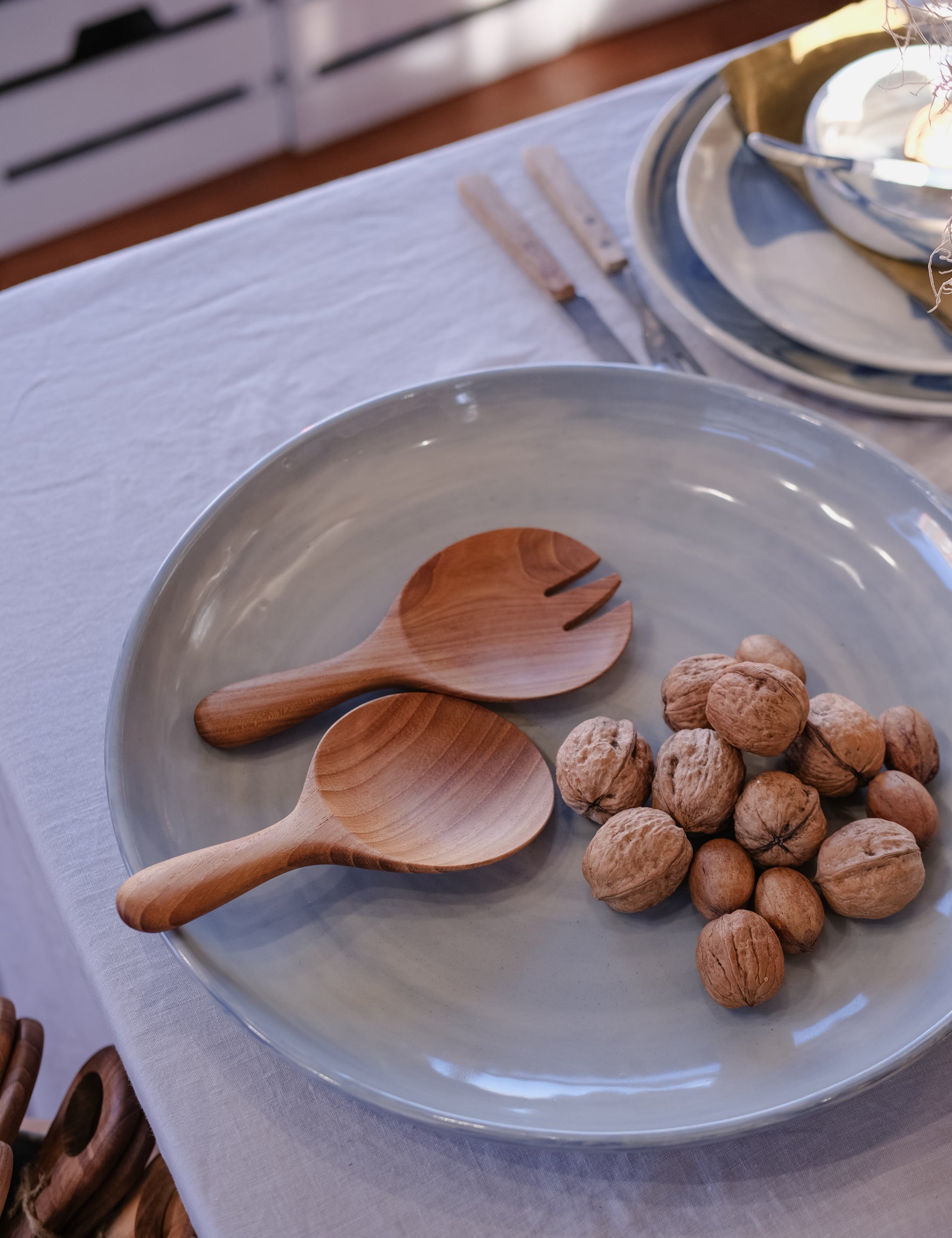 A light blue-gray ceramic plate on a grey tablecloth, styled with two wooden salad serving spoons and a small pile of walnuts. A matching stack of handmade ceramic plates and cutlery is visible in the softly blurred background.