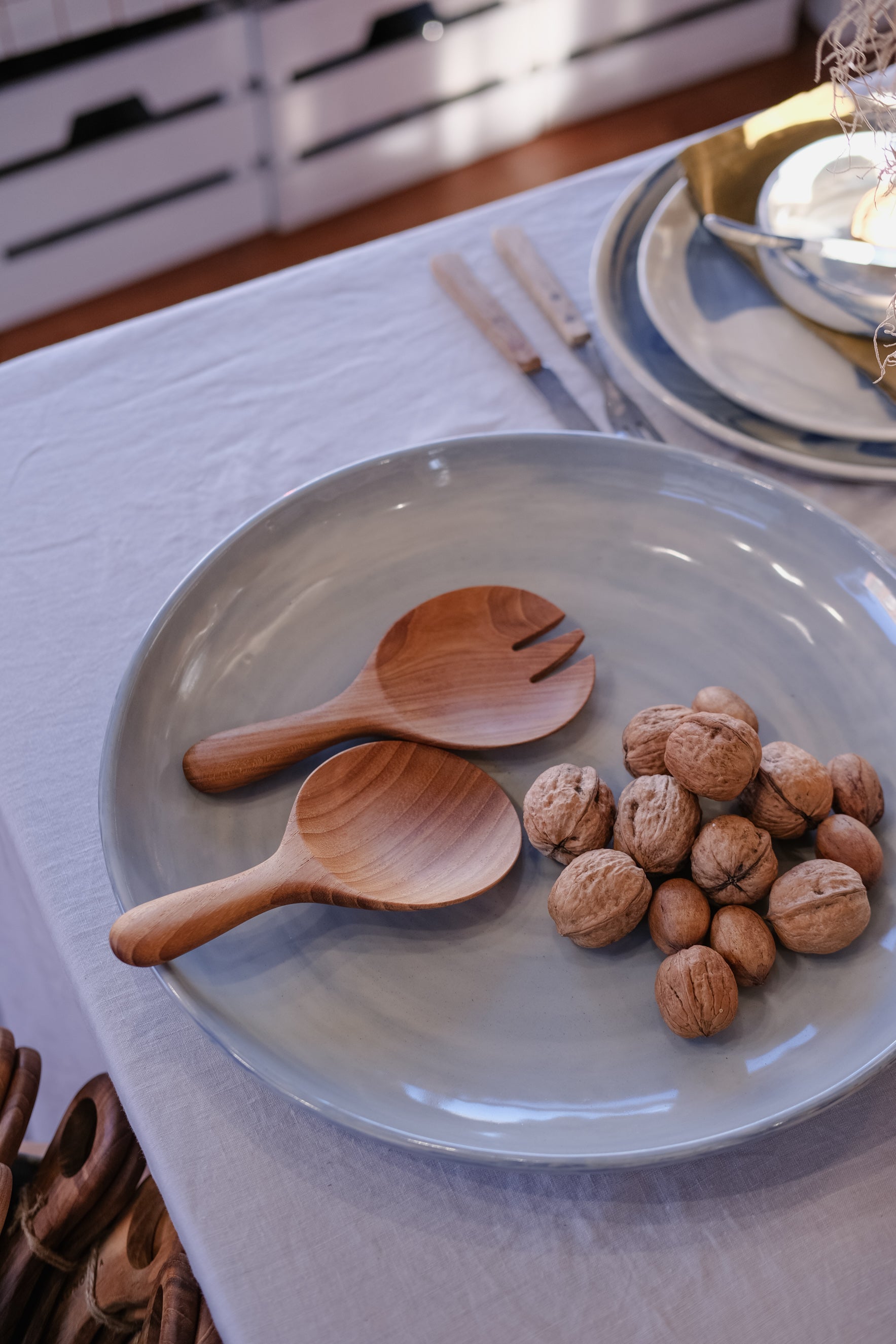A light blue-gray ceramic plate on a grey tablecloth, styled with two wooden salad serving spoons and a small pile of walnuts. A matching stack of handmade ceramic plates and cutlery is visible in the softly blurred background.