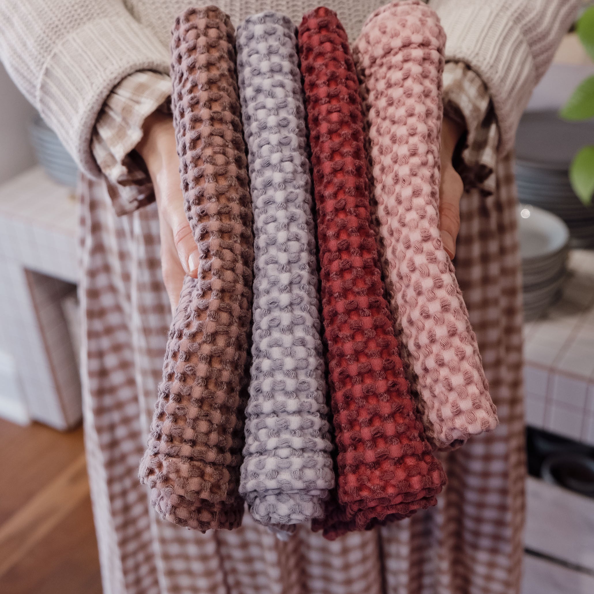 A person in a beige knit sweater and checkered skirt holds four textured waffle-weave tea towels in brown, grey, rust red, and dusty pink, shown against a warm indoor background.