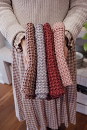 A person in a beige knit sweater and checkered skirt holds four textured waffle-weave tea towels in brown, grey, rust red, and dusty pink, shown against a warm indoor background.