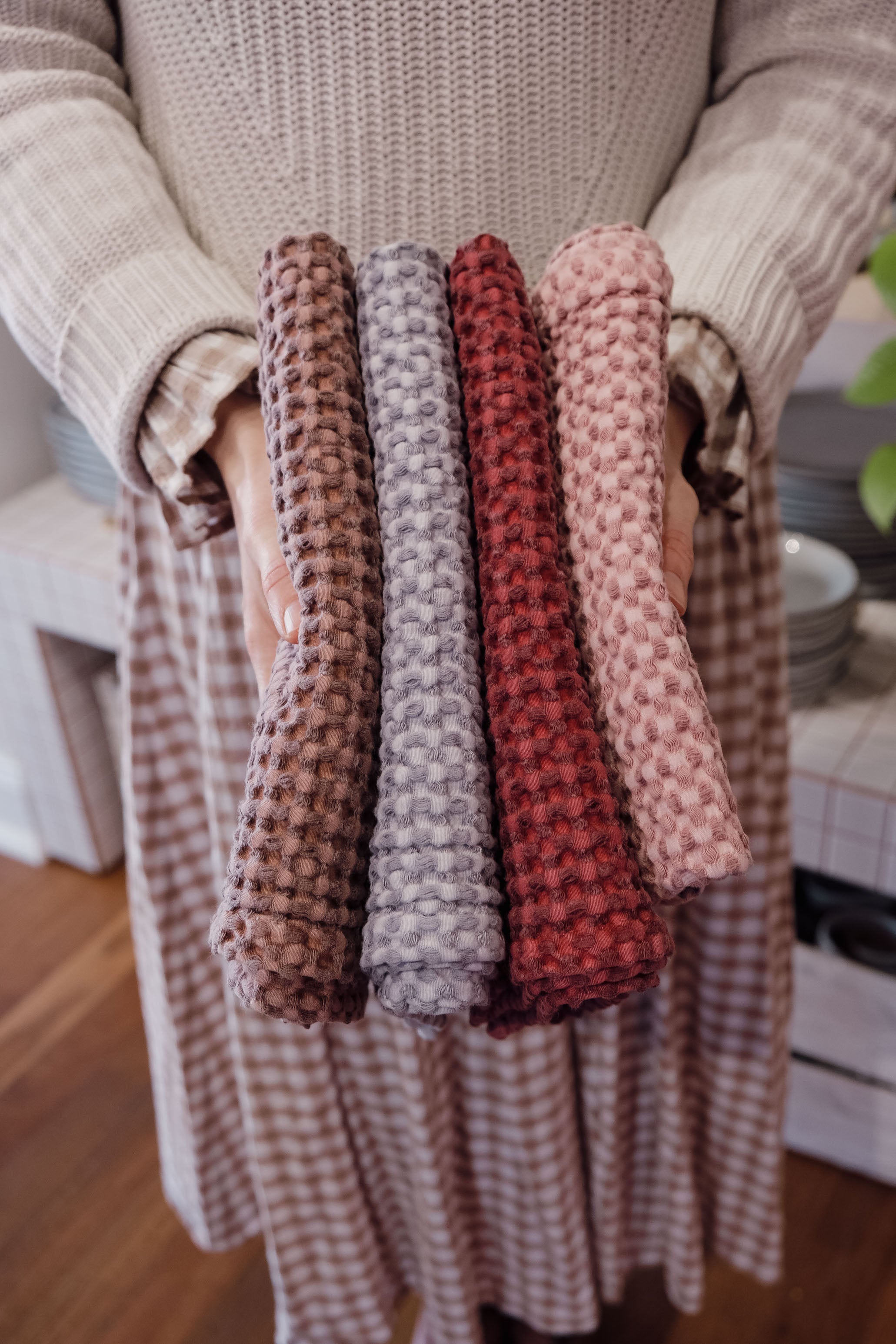A person in a beige knit sweater and checkered skirt holds four textured waffle-weave tea towels in brown, grey, rust red, and dusty pink, shown against a warm indoor background.