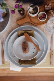 A styled tabletop setting featuring a speckled light blue ceramic three piece dinner set, bread plate, lunch plate and a dinner plate, and matching accessories, a teak wooden spoon, a folded olive green napkin, and rustic dining props including flowers, mushrooms, and a glass on a wooden board.