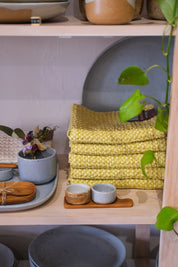 A shelf in a homewares store full of handmade blue speckled ceramics, wooden homewares and serving dishes and vintage yellow waffle towels stacked.
