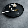 Side view of a Handmade black ceramic dinner plate by Palinopsia Ceramics  in a still life shot with garlic and salt